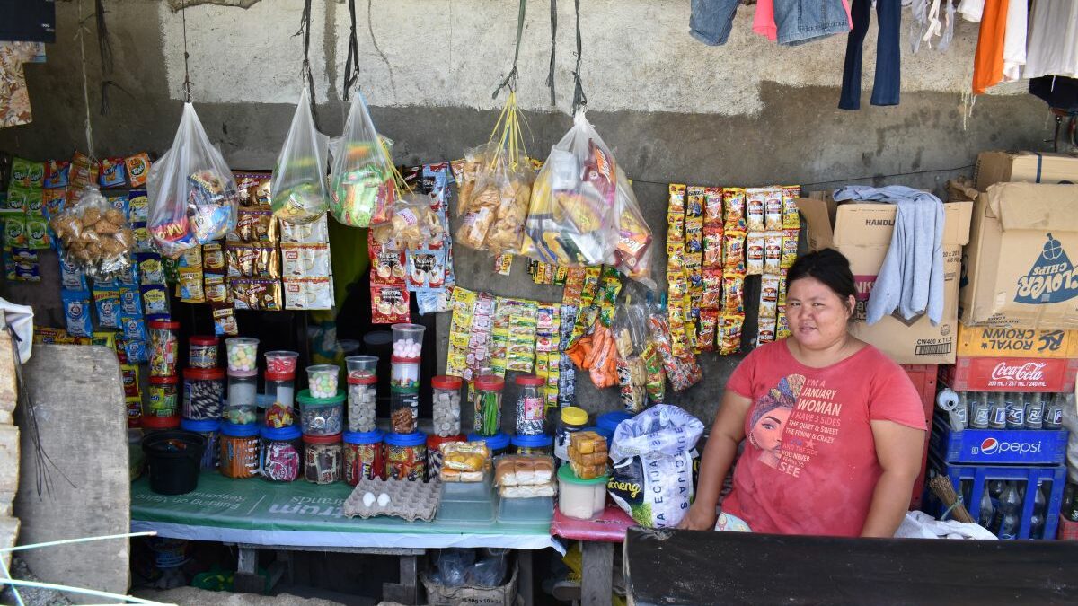 Carmela sitzt in ihrem kleinen Kiosk auf den Philippinen. An der Rückwand hängen bunte Waren, Lebensmittel stehen teilweise in Dosen auf dem Tisch neben ihr.