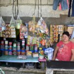 Carmela sitzt in ihrem kleinen Kiosk auf den Philippinen. An der Rückwand hängen bunte Waren, Lebensmittel stehen teilweise in Dosen auf dem Tisch neben ihr.