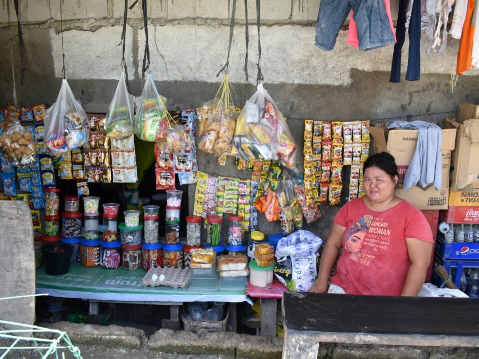 Carmela sitzt in ihrem kleinen Kiosk auf den Philippinen. An der Rückwand hängen bunte Waren, Lebensmittel stehen teilweise in Dosen auf dem Tisch neben ihr.