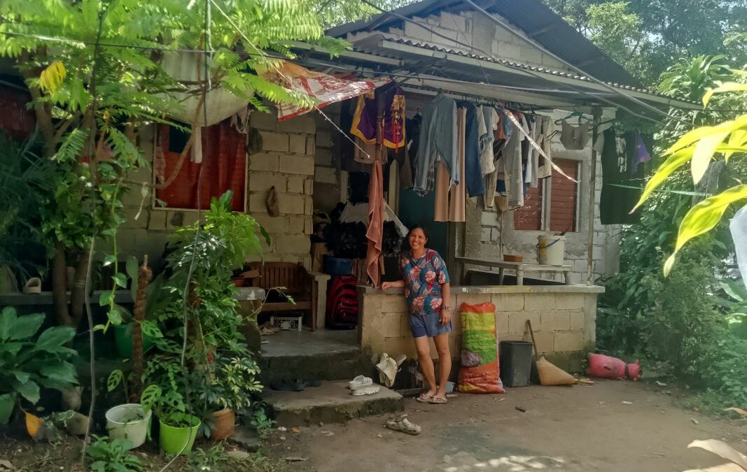 Sally steht barfuß auf festgetretenem Sandboden vor einfachem Haus mit unverputzter Steinwand, aufgehängter Kleidung, Pflanzen in Töpfen.