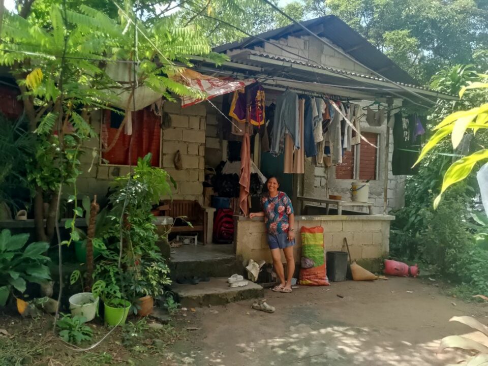 Sally steht barfuß auf festgetretenem Sandboden vor einfachem Haus mit unverputzter Steinwand, aufgehängter Kleidung, Pflanzen in Töpfen.