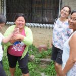 Vier philippinische Kleinstunternehmerinnen stehen lachend im Freien vor einem Haus mit vergittertem Fenster und genießen das Gespräch miteinander.