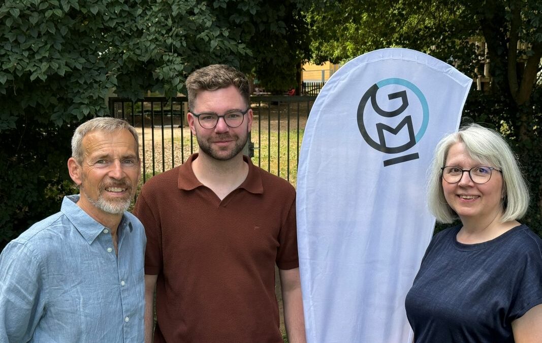 Die drei Vorstandsmitglieder von Global Micro Initiative e.V. stehen nebeneinander vor einem GMI-Banner im Freien und lächeln in die Kamera. Im Hintergrund Bäume und ein Zaun.