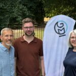 Die drei Vorstandsmitglieder von Global Micro Initiative e.V. stehen nebeneinander vor einem GMI-Banner im Freien und lächeln in die Kamera. Im Hintergrund Bäume und ein Zaun.