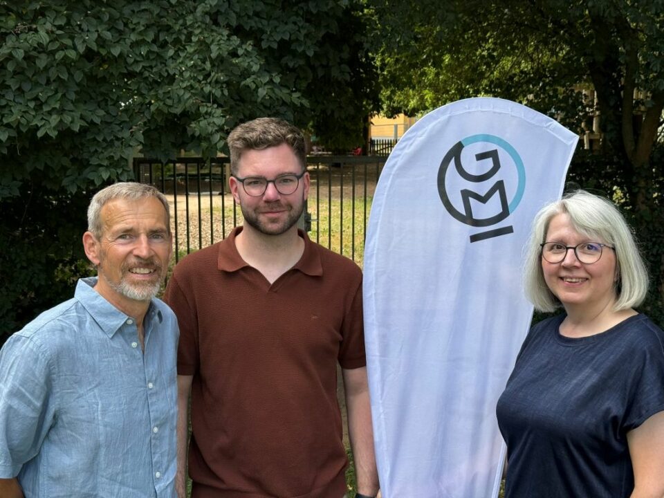 Die drei Vorstandsmitglieder von Global Micro Initiative e.V. stehen nebeneinander vor einem GMI-Banner im Freien und lächeln in die Kamera. Im Hintergrund Bäume und ein Zaun.