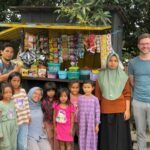 Gruppenbild vor einem kleinen Verkaufsstand in Jineng mit Kleinstunternehmerin, Kindern, Team von Global Micro Initiative e.V. und Tobias Schüßler.