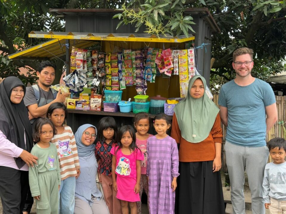 Gruppenbild vor einem kleinen Verkaufsstand in Jineng mit Kleinstunternehmerin, Kindern, Team von Global Micro Initiative e.V. und Tobias Schüßler.