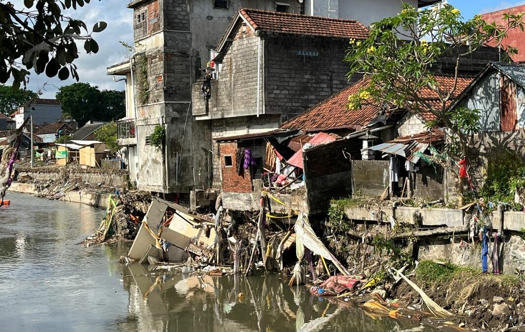 Wohnhäuser direkt am Flussufer in Denpasar, beschädigt durch Überschwemmung, Trümmer und Müll hängen an Mauern und Bäumen.