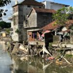 Wohnhäuser direkt am Flussufer in Denpasar, beschädigt durch Überschwemmung, Trümmer und Müll hängen an Mauern und Bäumen.