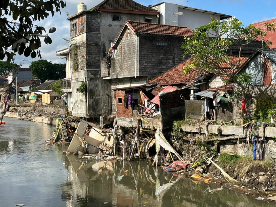 Wohnhäuser direkt am Flussufer in Denpasar, beschädigt durch Überschwemmung, Trümmer und Müll hängen an Mauern und Bäumen.