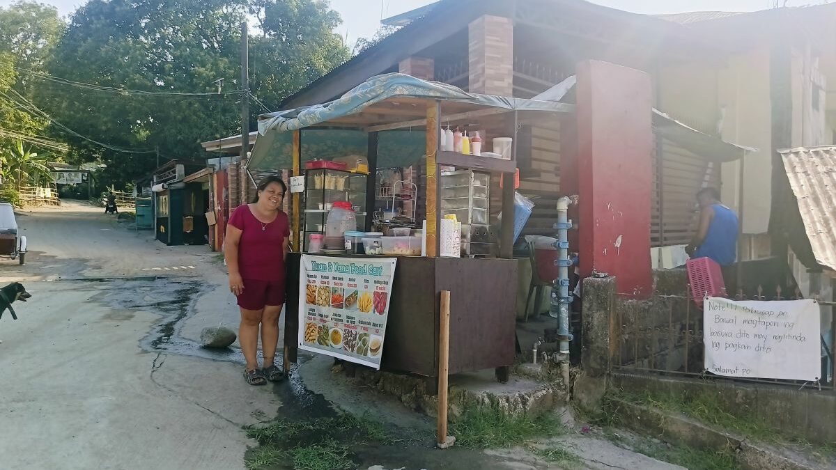 Eine Frau steht neben einem überdachten Straßenimbiss mit Auslage, Menütafel und Essensbehältern an einer Straße.