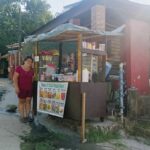 Eine Frau steht neben einem überdachten Straßenimbiss mit Auslage, Menütafel und Essensbehältern an einer Straße.
