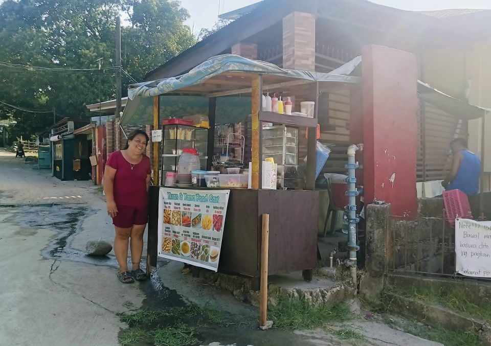 Eine Frau steht neben einem überdachten Straßenimbiss mit Auslage, Menütafel und Essensbehältern an einer Straße.
