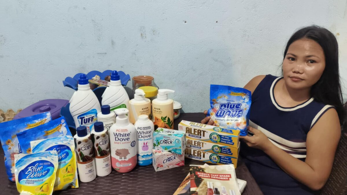 Ryla sitzt an einem Tisch mit verschiedenen Haushalts- und Körperpflegeprodukten, darunter Waschmittel, Lotionen und Seifen.