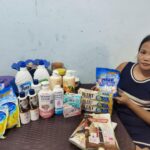 Ryla sitzt an einem Tisch mit verschiedenen Haushalts- und Körperpflegeprodukten, darunter Waschmittel, Lotionen und Seifen.