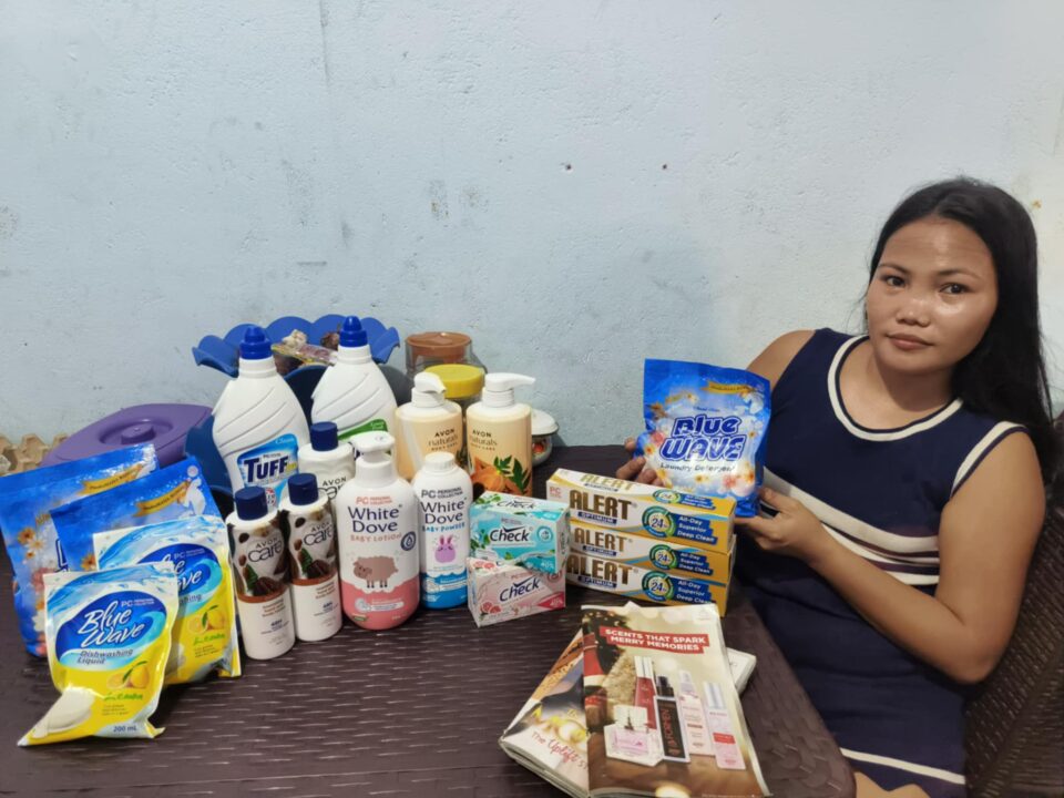 Ryla sitzt an einem Tisch mit verschiedenen Haushalts- und Körperpflegeprodukten, darunter Waschmittel, Lotionen und Seifen.