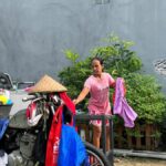 Aisyah steht in ihrem Hof mit einem Wäschestück in der Hand. Neben ihr hängen Kleidungsstücke zum Trocknen, im Vordergrund ein mit Wäsche beladenes Moped.