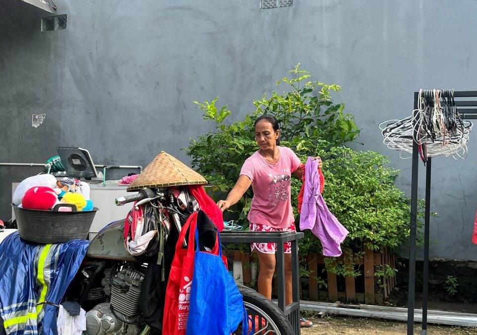 Aisyah steht in ihrem Hof mit einem Wäschestück in der Hand. Neben ihr hängen Kleidungsstücke zum Trocknen, im Vordergrund ein mit Wäsche beladenes Moped.
