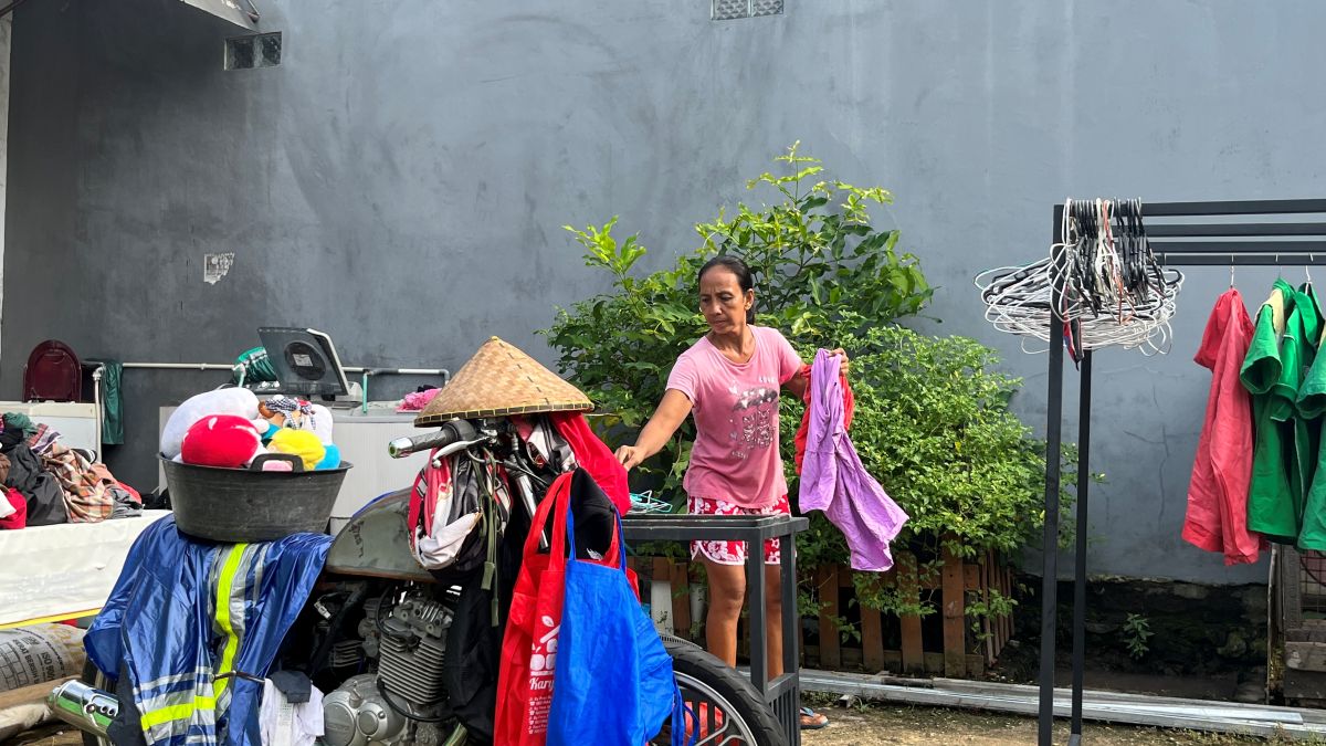 Aisyah steht in ihrem Hof mit einem Wäschestück in der Hand. Neben ihr hängen Kleidungsstücke zum Trocknen, im Vordergrund ein mit Wäsche beladenes Moped.