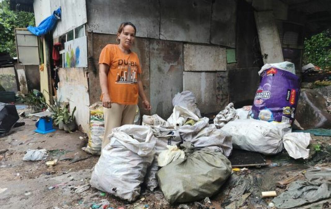 Merlin steht bei ihrem einfachen Haus aus Spanplatten, Wellblech und Planen. Neben ihr an der Wand lagern verschiedene gefüllte Säcke.
