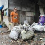 Merlin steht bei ihrem einfachen Haus aus Spanplatten, Wellblech und Planen. Neben ihr an der Wand lagern verschiedene gefüllte Säcke.