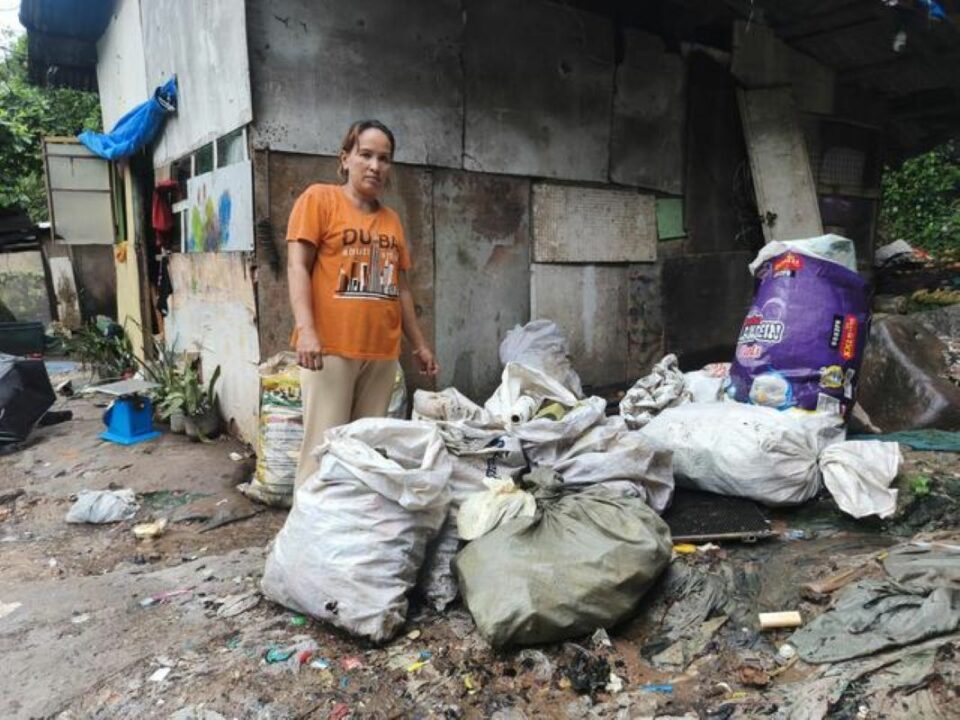 Merlin steht bei ihrem einfachen Haus aus Spanplatten, Wellblech und Planen. Neben ihr an der Wand lagern verschiedene gefüllte Säcke.