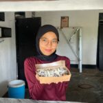 Mupidatul steht in ihrer einfachen Küche und hält eine Schachtel mit frisch zubereiteten Pisang Lumer zum Verkauf in die Kamera.