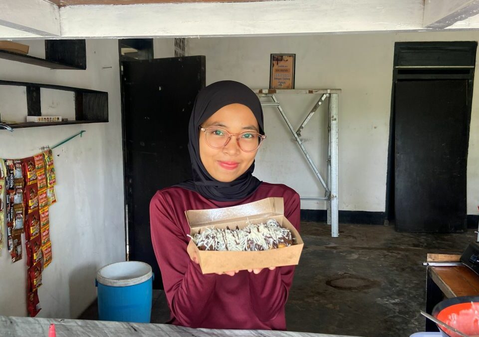 Mupidatul steht in ihrer einfachen Küche und hält eine Schachtel mit frisch zubereiteten Pisang Lumer zum Verkauf in die Kamera.