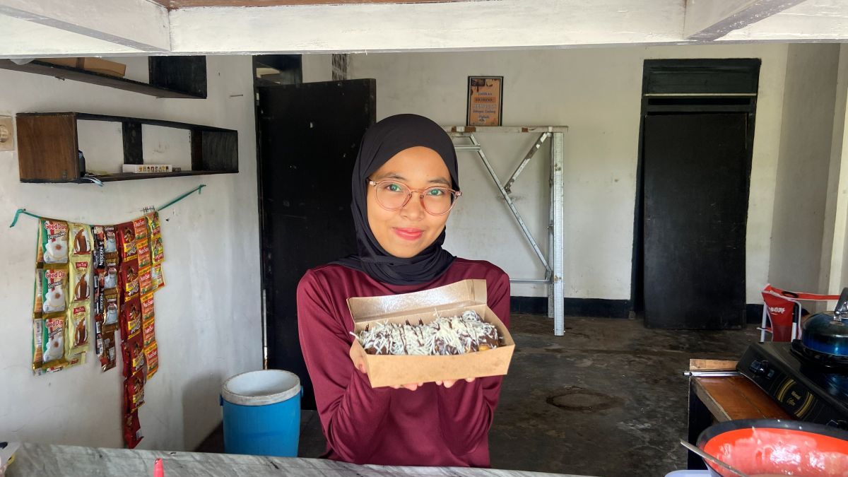 Mupidatul steht in ihrer einfachen Küche und hält eine Schachtel mit frisch zubereiteten Pisang Lumer zum Verkauf in die Kamera.