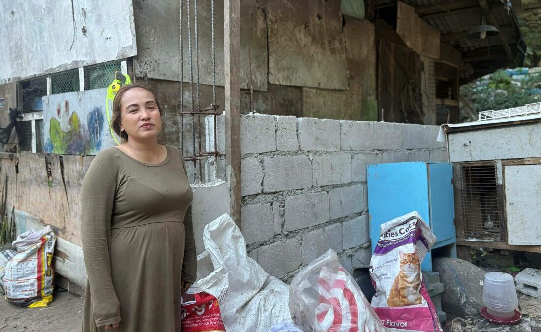 Frau steht vor teilweise gemauerter Hauswand in einer Siedlung nahe eines Müllbergs auf den Philippinen, mit Materialien für ihren Alltag im Vordergrund