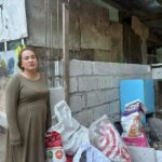 Frau steht vor teilweise gemauerter Hauswand in einer Siedlung nahe eines Müllbergs auf den Philippinen, mit Materialien für ihren Alltag im Vordergrund