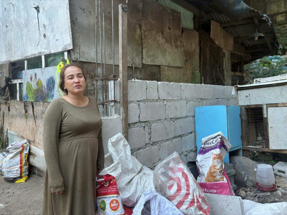 Frau steht vor teilweise gemauerter Hauswand in einer Siedlung nahe eines Müllbergs auf den Philippinen, mit Materialien für ihren Alltag im Vordergrund