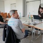 Die Vorstandsmitglieder Achim, Silvia und Tobias sitzen an einem großen Tisch. Vor Tobias steht ein aufgeklappter Laptop.