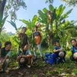 Die vier Mitglieder von GMI Philippines und zwei weitere Personen halten Kokospalmen-Setzlinge in den Händen. Zwei Personen stehen, die anderen sitzen in der Hocke. Im Hintergrund Bananenstauden und andere Bäume auf einem Hügel