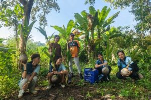 Die vier Mitglieder von GMI Philippines und zwei weitere Personen halten Kokospalmen-Setzlinge in den Händen. Zwei Personen stehen, die anderen sitzen in der Hocke. Im Hintergrund Bananenstauden und andere Bäume auf einem Hügel