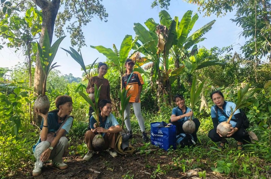 Die vier Mitglieder von GMI Philippines und zwei weitere Personen halten Kokospalmen-Setzlinge in den Händen. Zwei Personen stehen, die anderen sitzen in der Hocke. Im Hintergrund Bananenstauden und andere Bäume auf einem Hügel