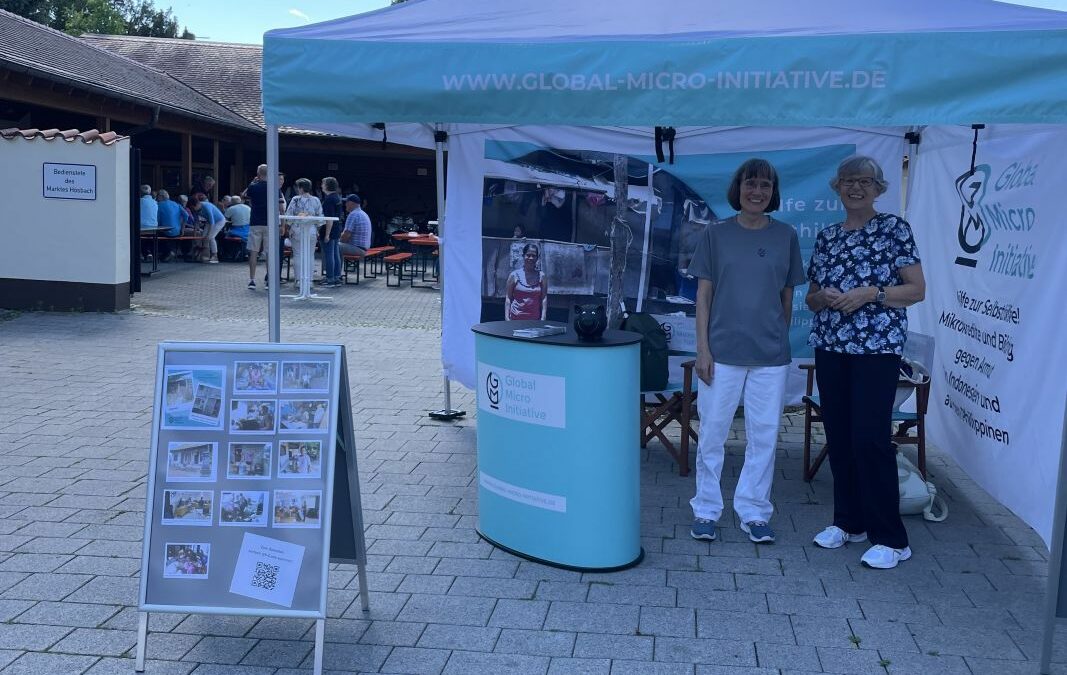Infostand der Global Micro Initiative mit Hanne und Eva‑Maria unter einem türkis‑weißen Pavillon; daneben eine Fototafel mit Bildern und QR‑Code, im Hintergrund sitzen Menschen and Festbank-Garnituren im Schatten eines Gebäudes.
