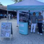 Infostand der Global Micro Initiative mit Hanne und Eva‑Maria unter einem türkis‑weißen Pavillon; daneben eine Fototafel mit Bildern und QR‑Code, im Hintergrund sitzen Menschen and Festbank-Garnituren im Schatten eines Gebäudes.