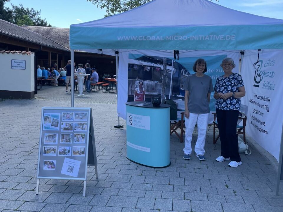 Infostand der Global Micro Initiative mit Hanne und Eva‑Maria unter einem türkis‑weißen Pavillon; daneben eine Fototafel mit Bildern und QR‑Code, im Hintergrund sitzen Menschen and Festbank-Garnituren im Schatten eines Gebäudes.