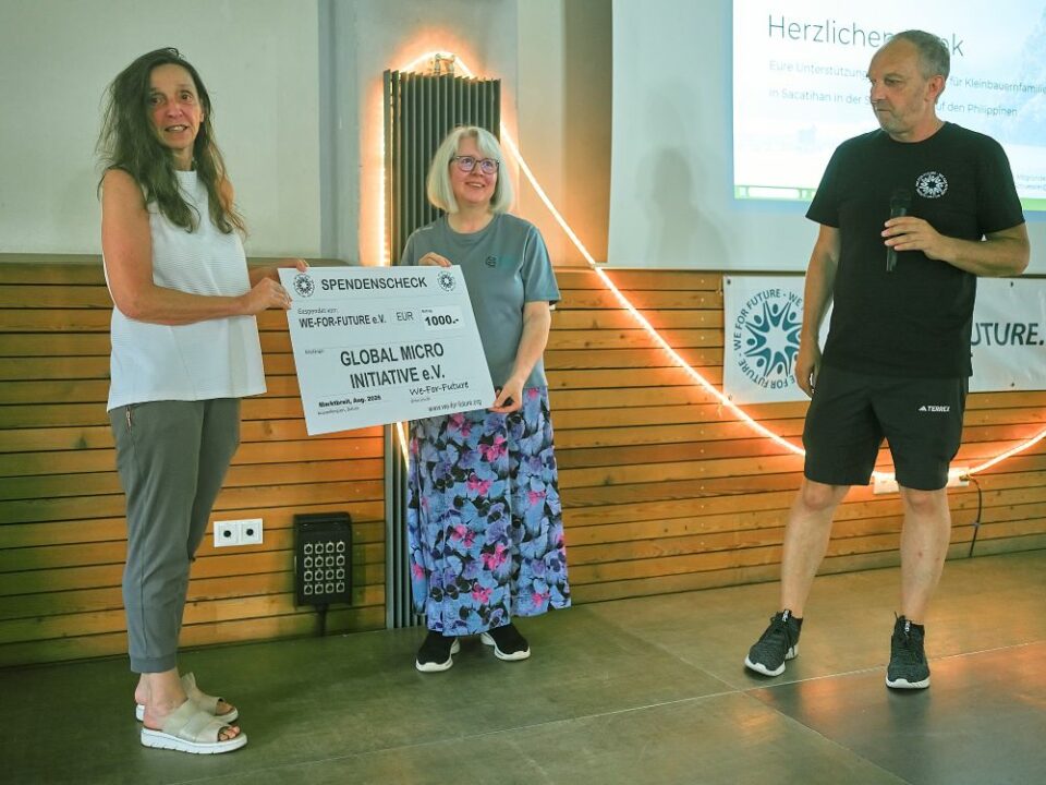 Eine Frau überreicht Silvia von Global Micro Initiative e.V. einen Spendenscheck über 1.000 Euro von WE-FOR-FUTURE e. V.